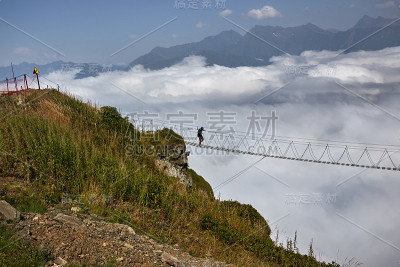 人们走在吊桥上，看着下面多云的山。