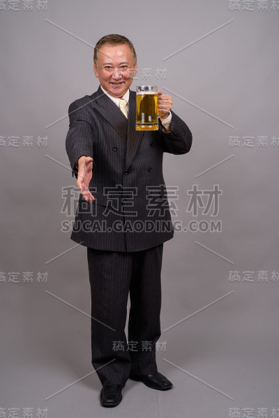 在灰色的背景下，一个成熟的亚洲商人拿着一杯啤酒