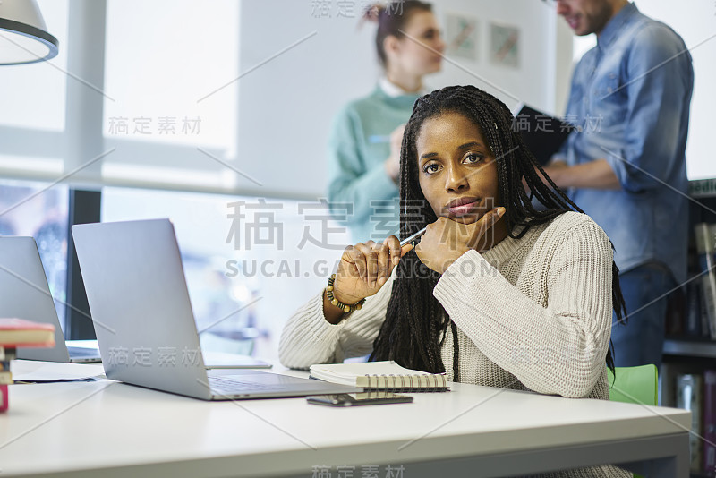 这是一幅深思熟虑的年轻美国黑人国际学生坐在大学图书馆的桌子旁，用笔记本电脑和无线网络做作业，而同学们