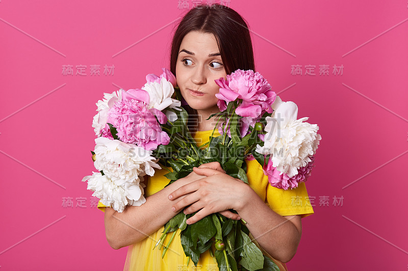 近距离肖像的铅笔深色头发的女人拥抱完美的牡丹花，摆出孤立在玫瑰色的背景，女性看一边与体贴和狡猾的面部