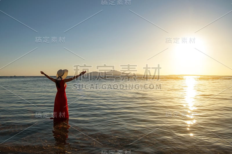 初夏清晨，一名身穿红色长裙、头戴草帽的年轻女子站在海边的海水中，举手欣赏初升的太阳。