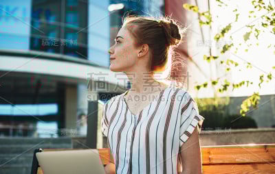 迷人的红头发白种人女孩与雀斑看着别处，而坐在一个长凳在城市