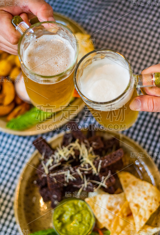 近距离观看两杯啤酒在手。酒杯在酒吧或酒吧的桌子上叮当作响，以食物为背景