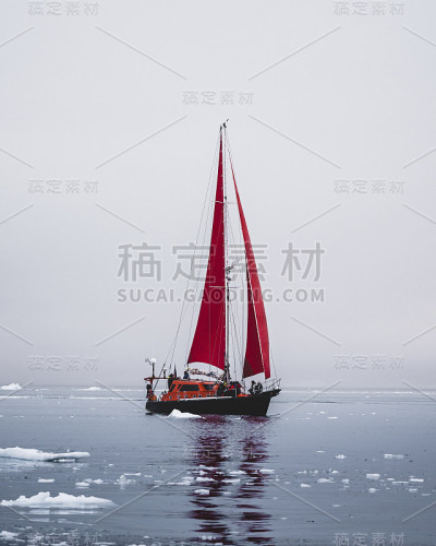 美丽的红色帆船在北极附近的巨大冰山显示规模。在极地夏季的午夜太阳季节，在迪斯科湾冰川的浮冰之间游弋。