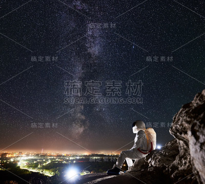 宇航员坐在布满星星的壮丽夜空下的岩石山上。