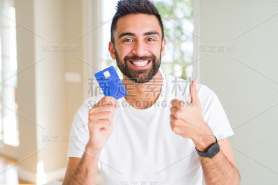 英俊的西班牙男人拿着信用卡开心地笑着做ok的手势，大拇指向上的手指，优秀的手势