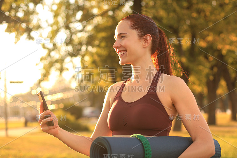 可爱微笑的女人穿着运动服使用智能手机，在日落公园拿着瑜伽垫。