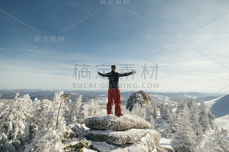 一名徒步者举起双手，站在悬崖顶上，欣赏冬季的山景。快乐的自由滑雪板游客在冬天