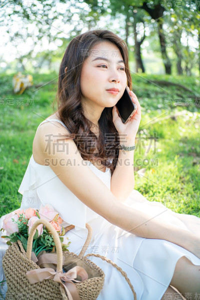 迷人的亚洲美女坐在绿色草地上谈论智能手机