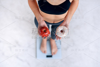 身材完美的运动女性拿着一个甜甜圈和一个红苹果用电子秤测量体重。