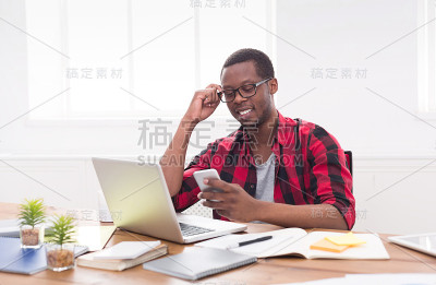 年轻的非洲裔美国商人一边用笔记本电脑工作一边用手机发短信
