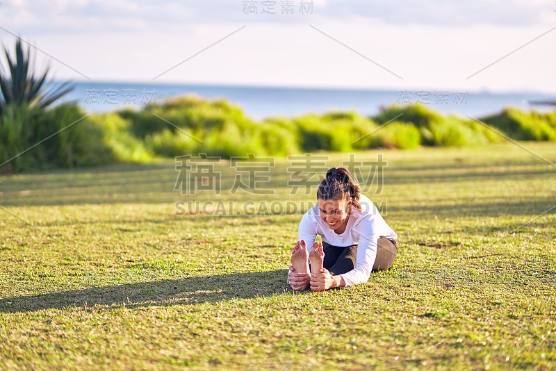 年轻美丽的女运动员微笑着练习瑜伽。教练面带微笑坐在公园里教坐前折叠姿势