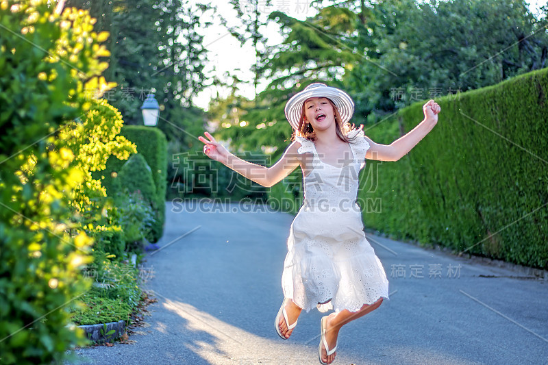 有趣的快乐的十岁前的女孩在白色的夏季连衣裙和帽子在户外街道上跳跃在阳光明媚的一天。现代活跃的疯狂少年