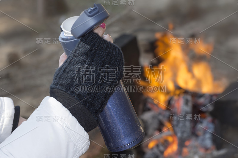 一个女人从篝火旁的保温瓶里喝咖啡。带盖的杯子，旅行热杯，可重复使用的热饮杯。