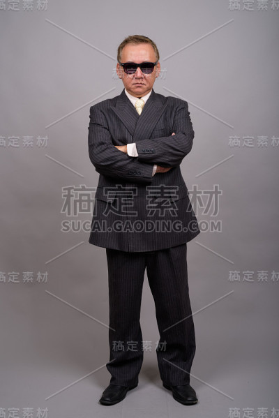 在灰色的背景下，成熟的亚洲商人戴着太阳镜看起来很酷