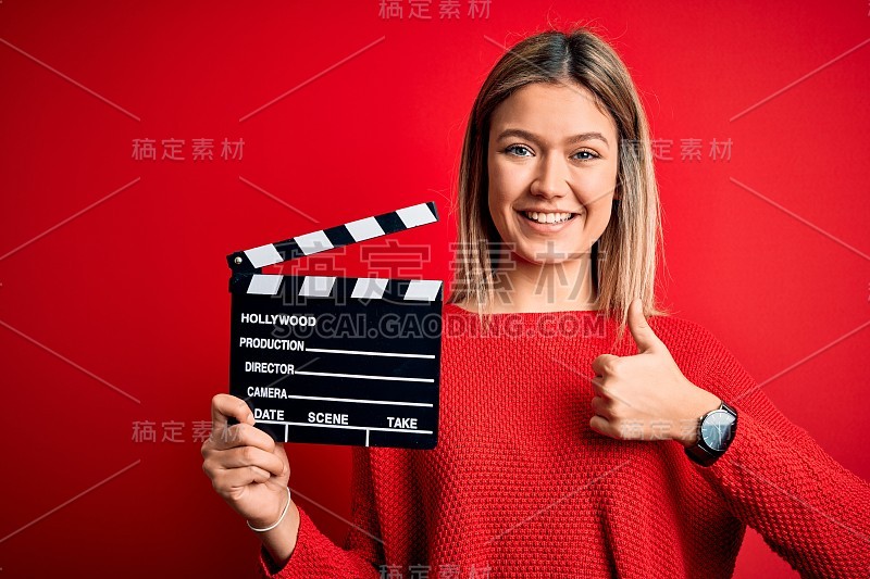 年轻漂亮的女人拿着隔板站在孤立的红色背景愉快地笑着做ok的手势，大拇指向上的手指，优秀的手势