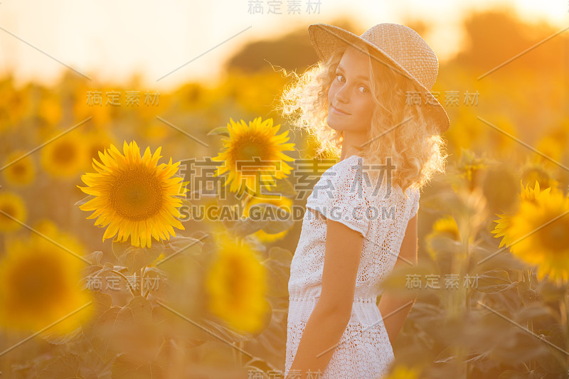 美丽的卷发女孩穿着白色的裙子，在夕阳下的向日葵田野