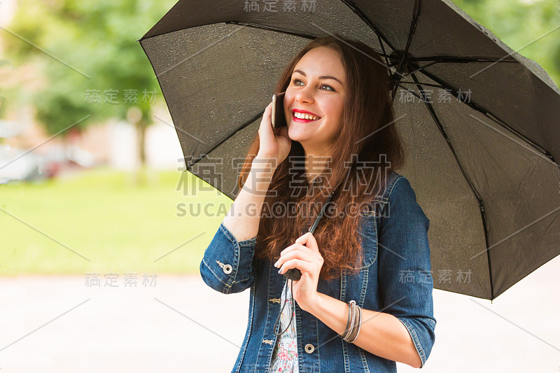 年轻女子撑着伞，在雨天在户外打手机