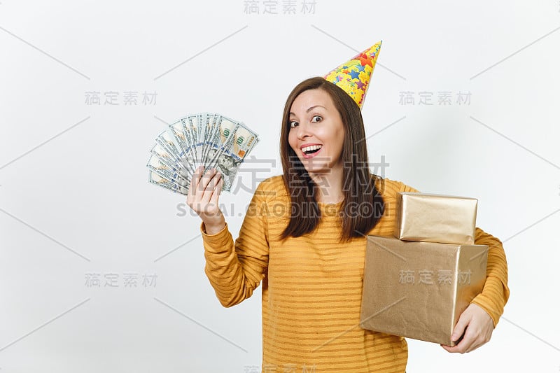 白种人有趣的年轻快乐的女人在黄色的衣服，生日派对帽子拿着金礼盒与礼物和大把的现金，庆祝节日在白色的背