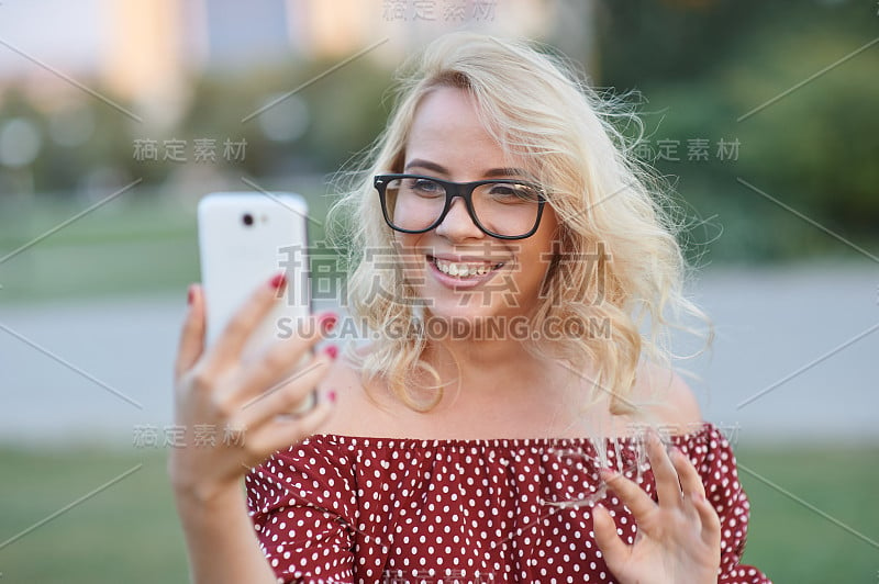 美丽的年轻女子金发博主在城市街道的背景下用智能手机拍照。有智能手机的漂亮女孩