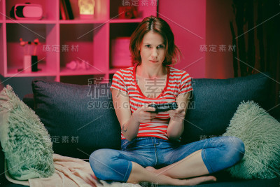 美丽兴奋的年轻玩家女孩坐在沙发上，在主机上玩视频游戏。她玩无线控制器。温馨的房间，以温暖和霓虹灯照明