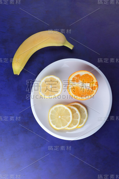 极简主义风格的紫外光背景柑橘，白色盘子上的柠檬片和半片橙子，深蓝色背景上的黄色香蕉，健康食品，素食，