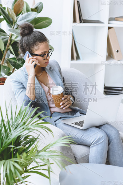 非裔美国女商人坐在扶手椅上一边用笔记本电脑工作一边用智能手机聊天