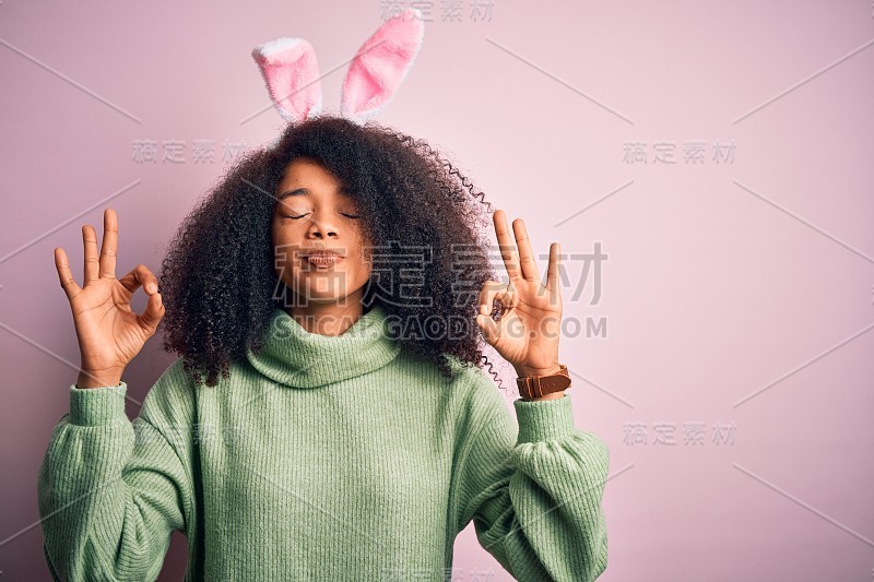 年轻的非洲裔美国妇女与非洲发式穿复活节兔子耳朵服装在粉红色的背景放松和微笑闭上眼睛做冥想手势与手指。