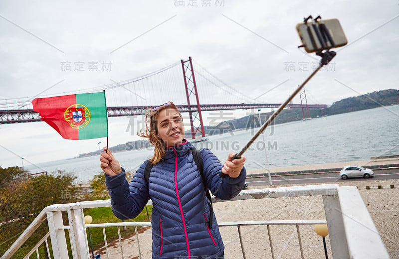 年轻女子游客自拍在著名的铁4月25日桥的背景和手握葡萄牙国旗在里斯本市