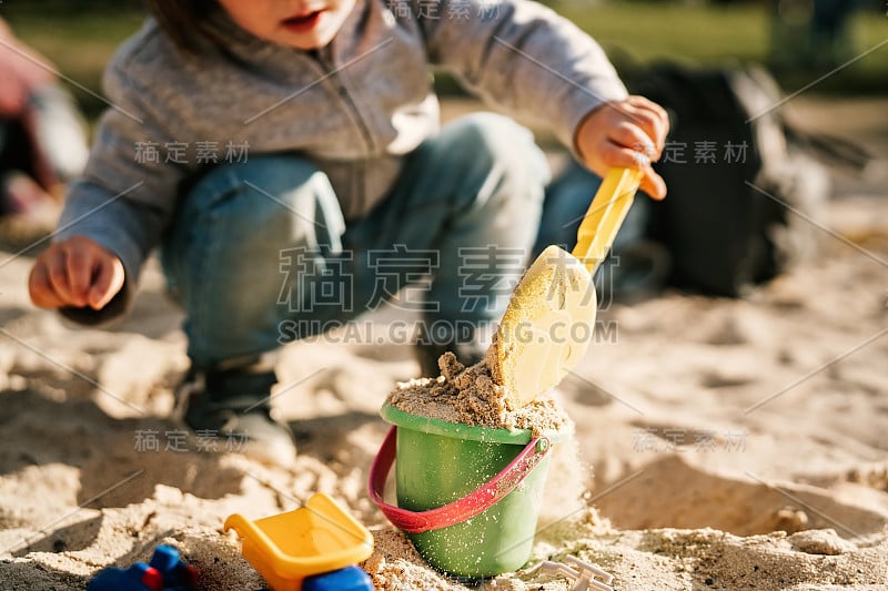 小孩子在公园外面玩沙子，用铲子把沙子装进桶里。选择性地关注沙子。户外休闲活动。学习通过玩耍