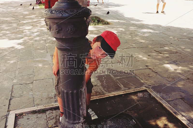 在一个炎热的夏天，美丽的淘气男孩在威尼斯广场的饮水机里喝水