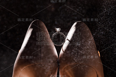 一双白色鞋子之间有一枚金色的钻石戒指，背景是黑色