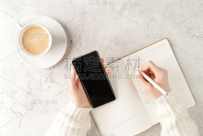 美丽女人的手拿着最新的智能手机，在笔记本上写着，在混凝土背景下喝着咖啡。妇女的经营理念。咖啡休息顶视