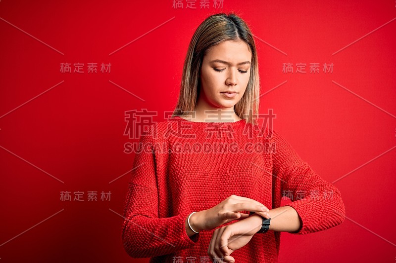 年轻美丽的金发女人穿着休闲毛衣越过红色孤立的背景检查手表上的时间，轻松和自信