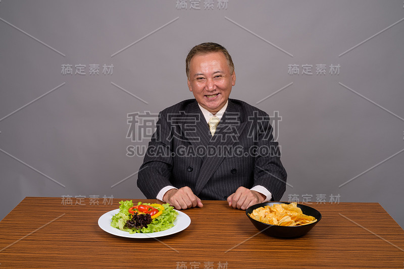 工作室拍摄的成熟的亚洲商人坐在木桌子上，以灰色的背景