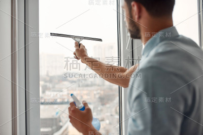 专家的工作。年轻的白人男子在客厅工作时，穿着制服用橡皮擦和喷雾清洁剂清洁窗户