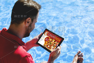 一名男子坐在泳池边，手里拿着平板电脑，屏幕上显示着一家披萨店的网站。