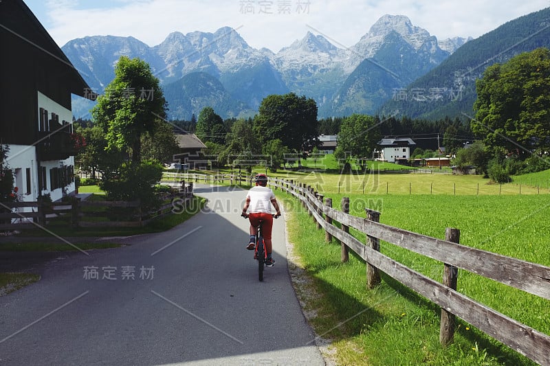 一个白人少年在奥地利的村庄街道上骑自行车，山上有绿色的草地在一个阳光明媚的夏天在他的假期