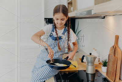 小女孩扎着辫子，围着围裙，学着做饭，在火炉旁摆姿势，准备早餐煎蛋，帮助父母做饭，在现代厨房里忙碌。儿