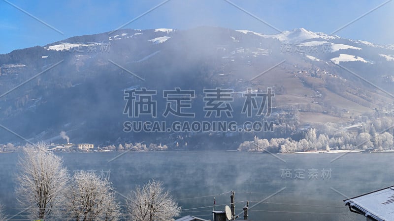 冬季明亮的奥地利阿尔卑斯山山坡上有阳光明媚的村庄房屋，雪和森林在雾蒙蒙的山岸边的湖