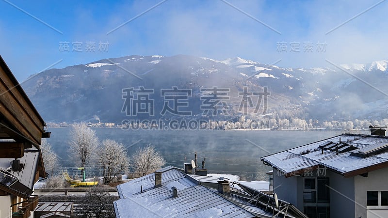 冬季明亮的奥地利阿尔卑斯山山坡上有阳光明媚的村庄房屋，雪和森林在雾蒙蒙的山岸边的湖