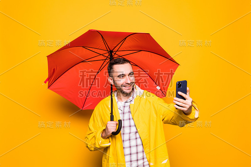 年轻英俊的胡子男子在黄色雨衣与红色伞自拍手机隔离在橙色背景