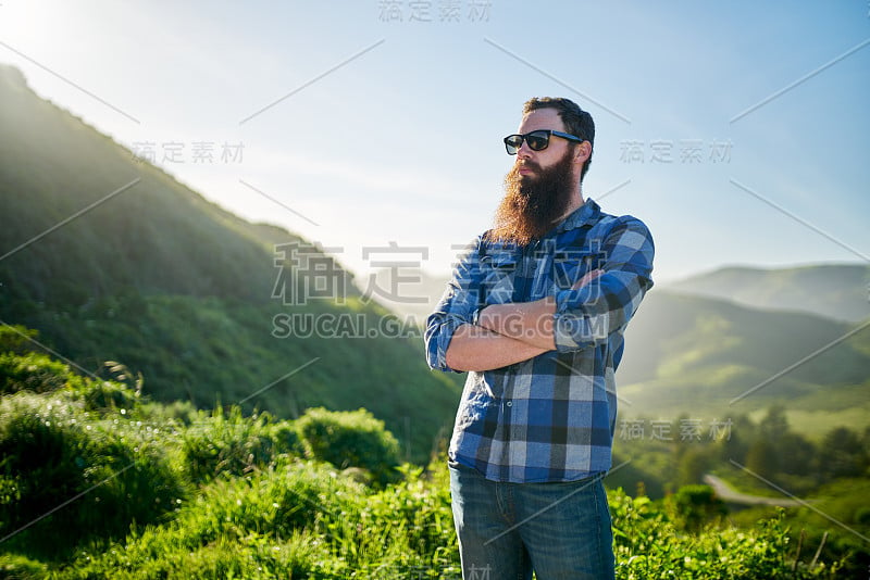 酷酷的蓄着胡子的家伙，戴着太阳镜，穿着蓝色法兰绒衣服，在绿色的草山旁摆姿势