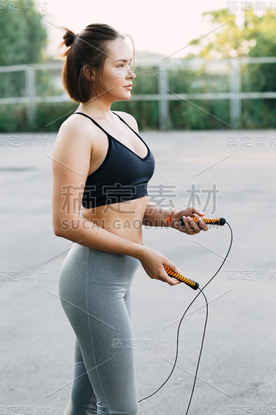 年轻迷人的女孩穿着运动服在操场上做运动，在夏天日落时跳绳