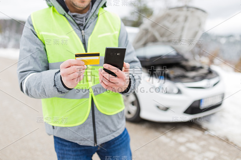 司机持有道路辅助服务会员卡和智能手机。背景是一辆开着引擎盖的破车。