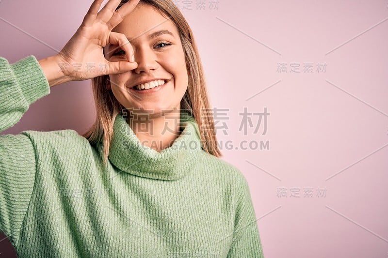 年轻美丽的金发女人穿着冬季羊毛衫在粉红色孤立的背景做ok手势与手微笑，眼睛透过手指与幸福的脸。