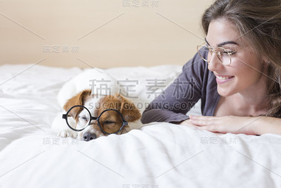 漂亮的年轻女子躺在床上，旁边还有她可爱的小狗。家，室内和生活方式。他们戴着时髦的眼镜