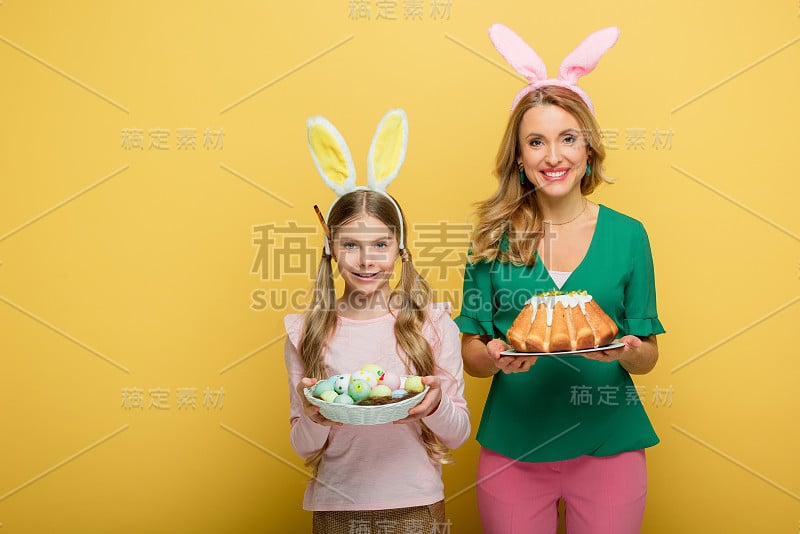 快乐的妈妈用兔耳朵抱着复活节蛋糕，旁边的女儿用彩绘的鸡蛋孤立在黄色的