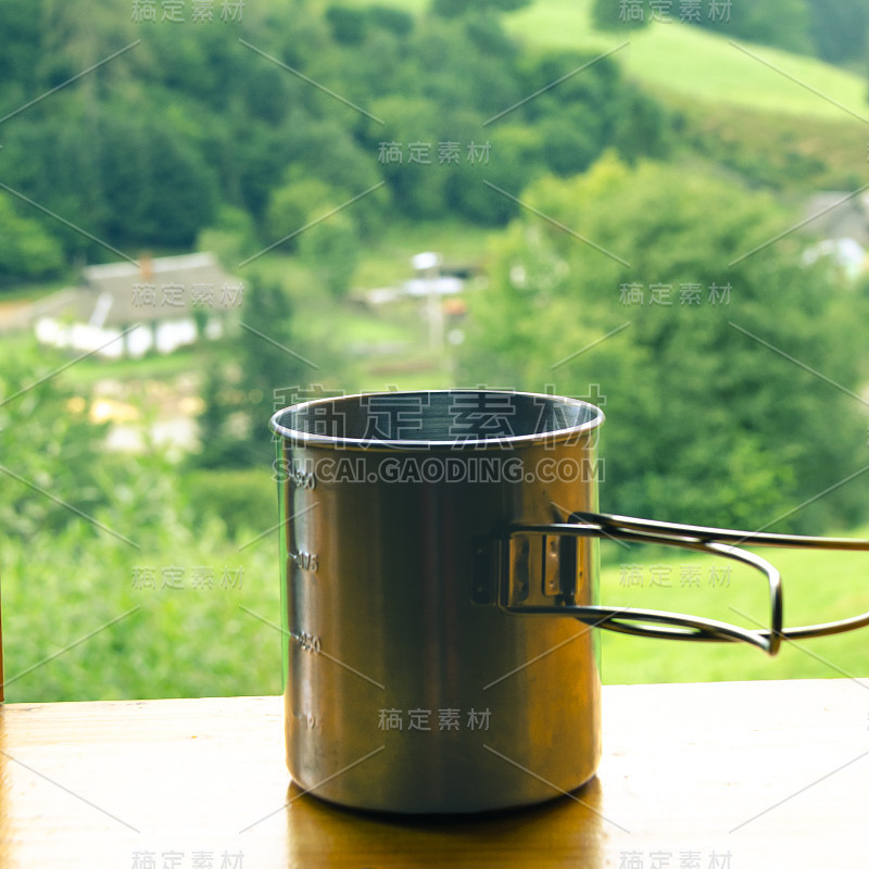 木窗台上放着一只金属茶杯，背景是群山