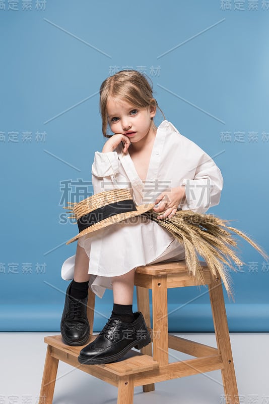 一个忧郁可爱的小女孩穿着白色的裙子，手拿麦穗和草帽，在蓝色的工作室拍摄